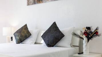 Un letto rifatto con cuscini a motivi, un comodino bianco con un vaso di fiori e un quadro appeso al muro.