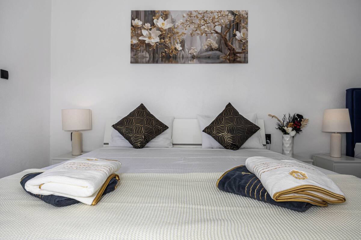 Una cama bien hecha con toallas, un cuadro sobre la cama y dos mesitas de noche con lámparas y un florero.