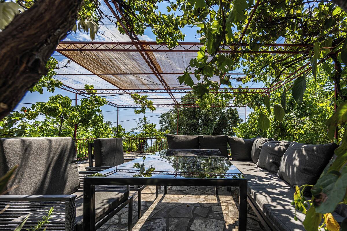 Un espacio de estar al aire libre con mesa de vidrio y sofás acolchados, rodeado de vegetación y una estructura metálica.