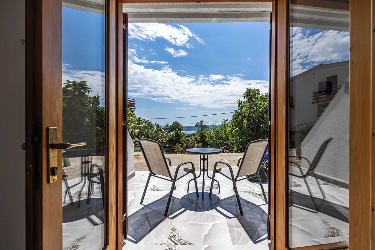 Puerta de vidrio que da a un patio exterior con mesa y sillas, vistas del cielo y algo de vegetación.