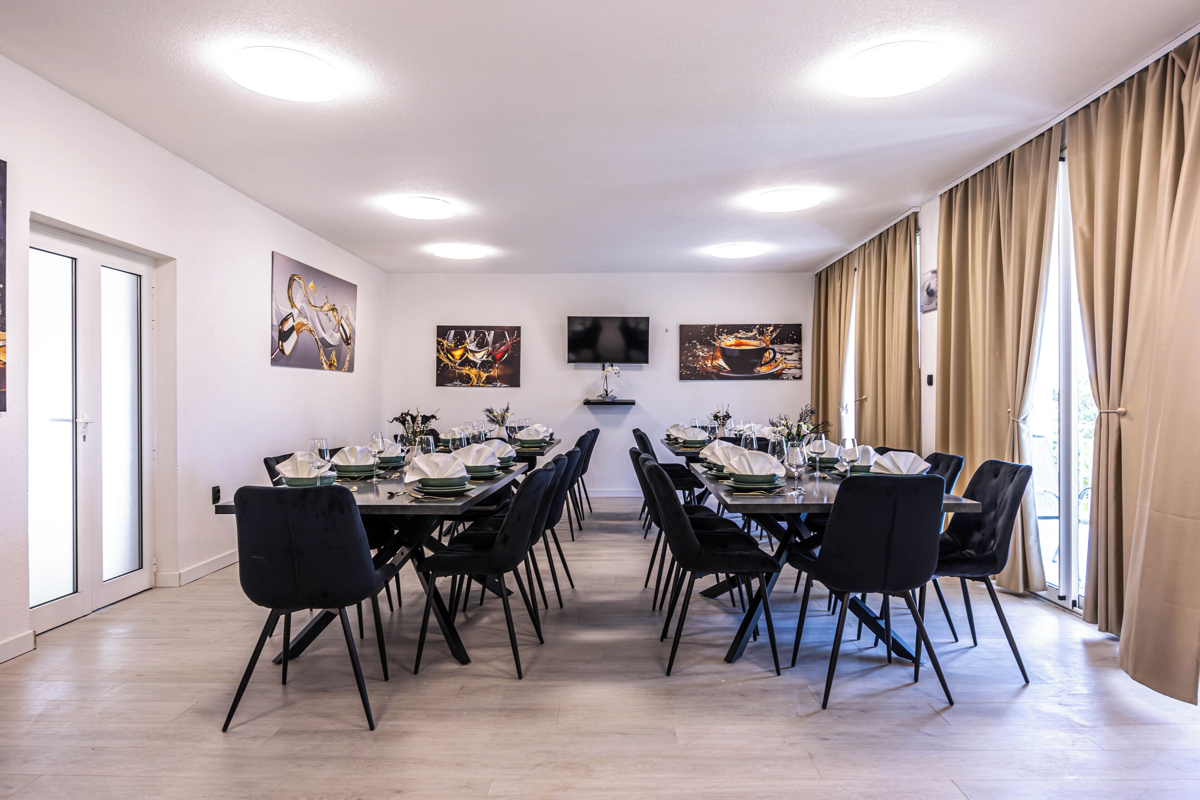 A dining room with a long table set for a meal, flanked by black chairs, with artwork on the walls and a flat-screen TV above.