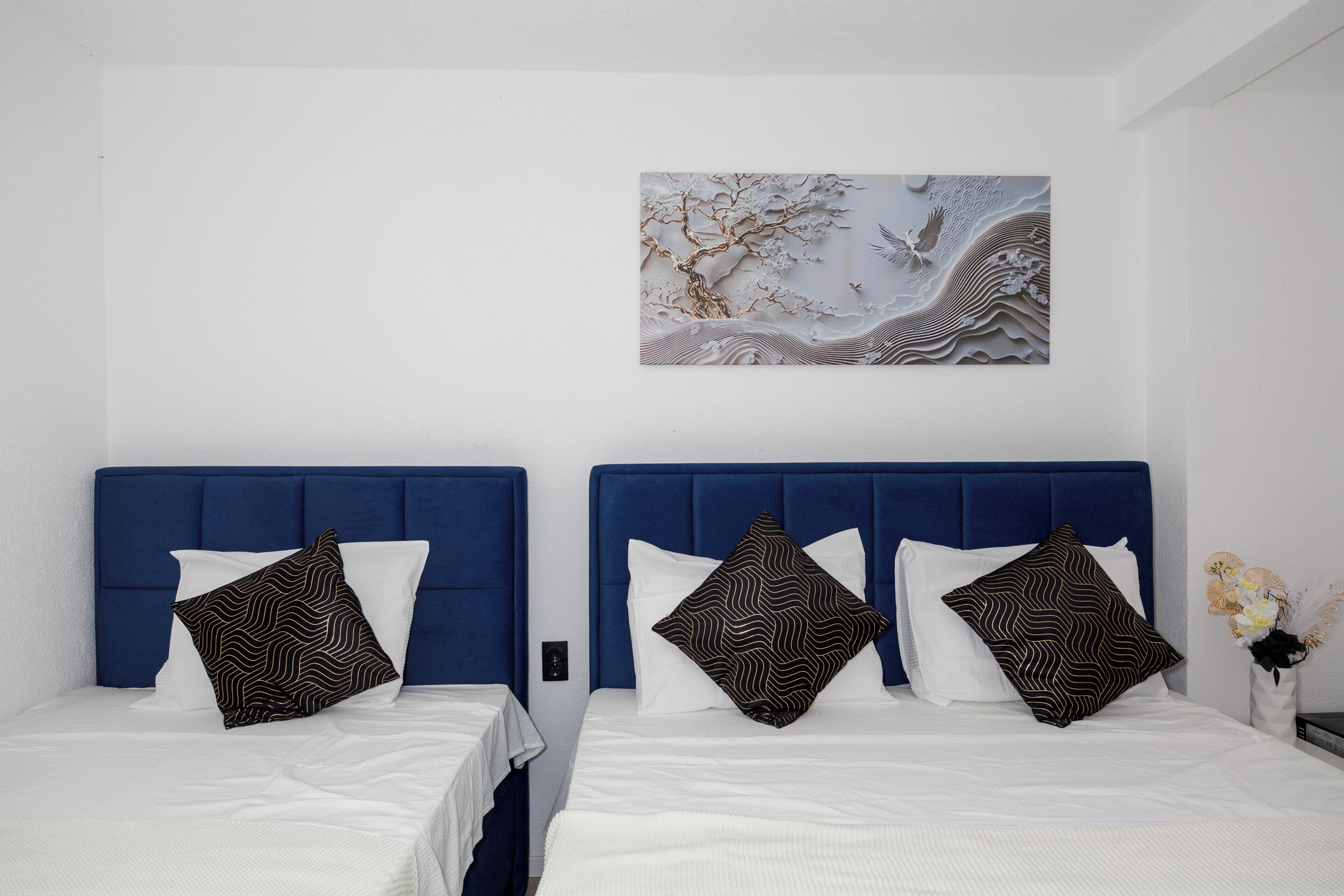 Ett hotellrum med två sängar, var och en med en blå sänggavel, vita sängkläder och mönstrade kuddar. Ett dekorativt väggobjekt hänger ovanför sängarna.