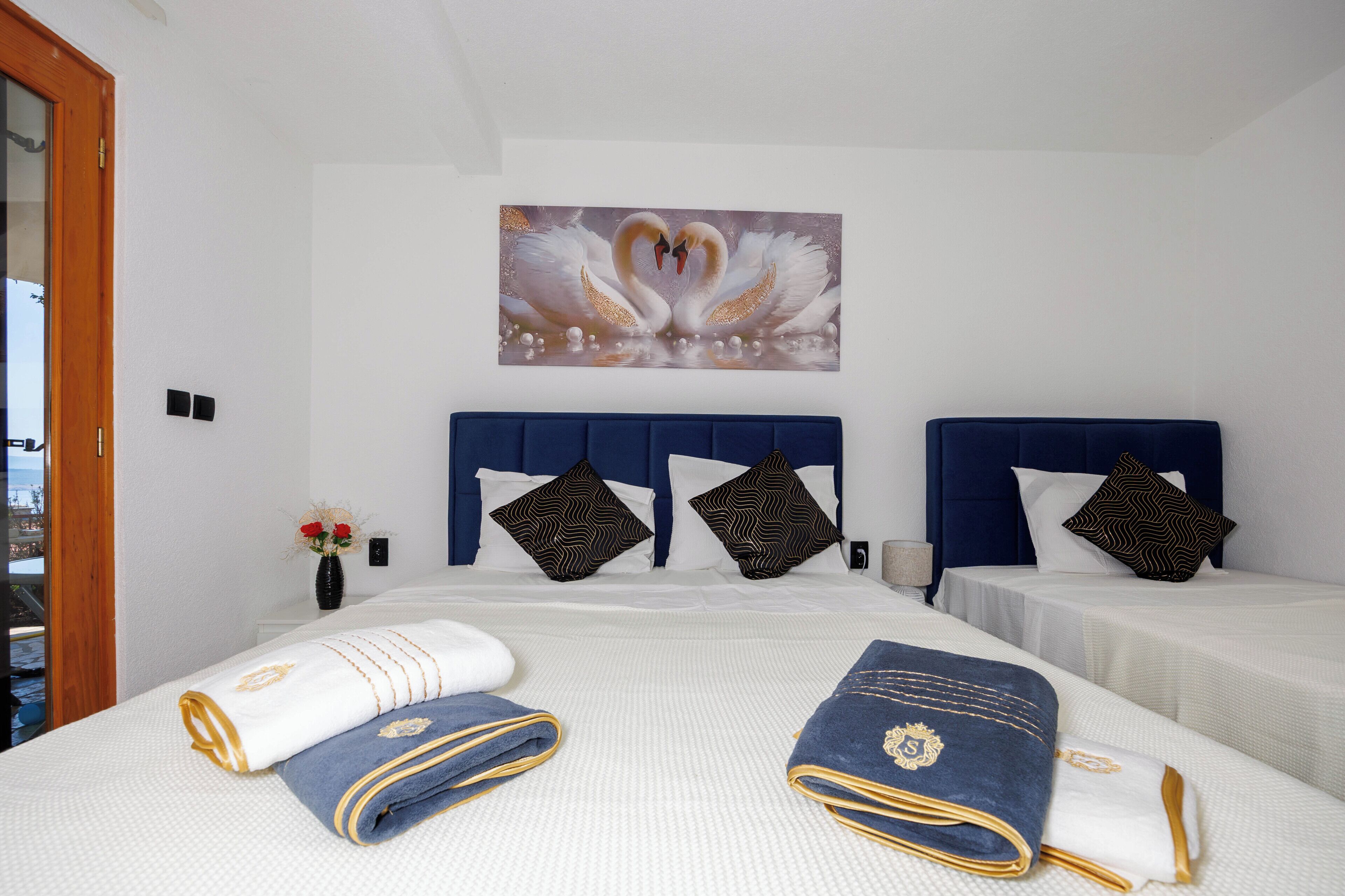 A hotel room with a large bed, two smaller beds, a painting of swans, and a vase with flowers.