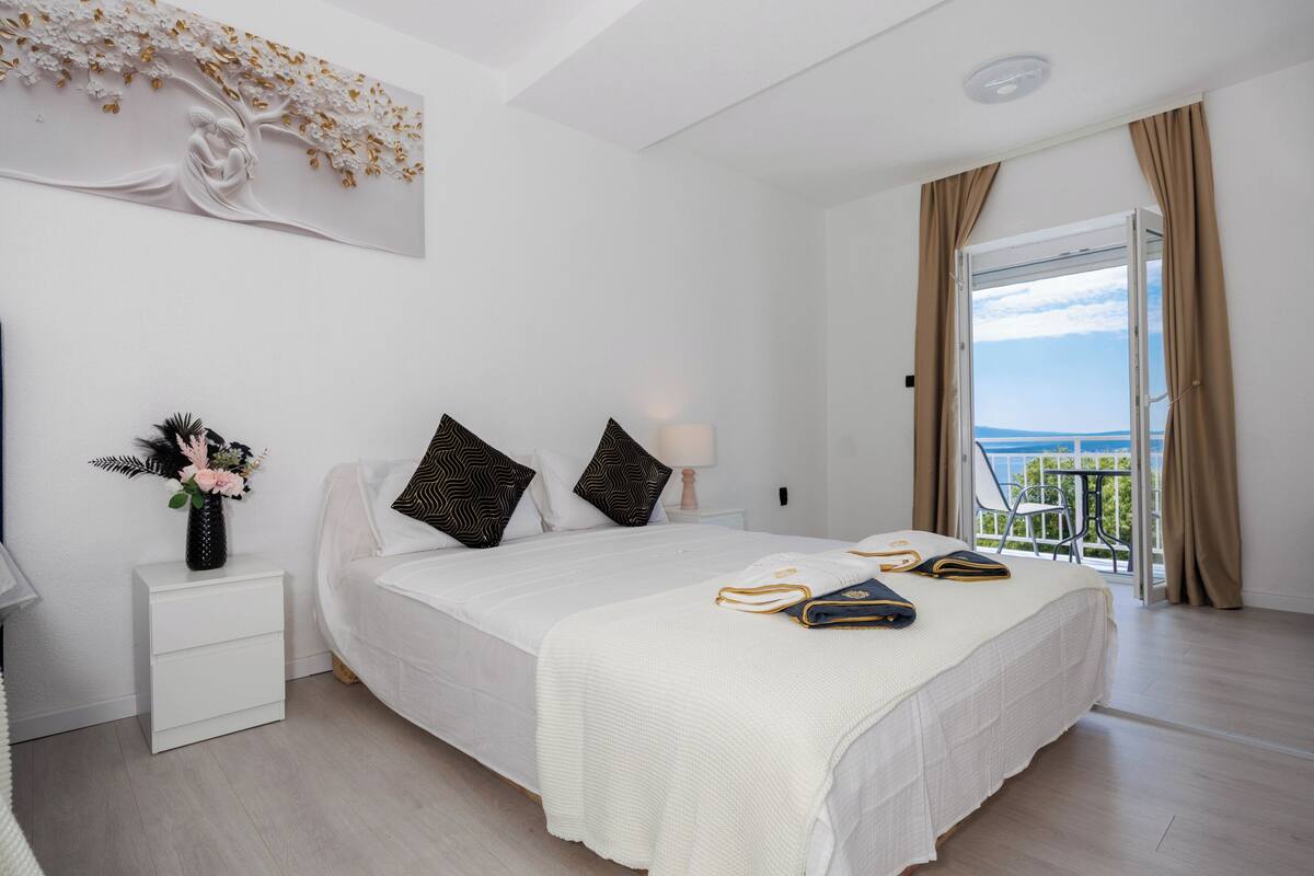 Un dormitorio con cama, mesita de noche, un jarrón con flores, ventana con cortinas y vistas al exterior.