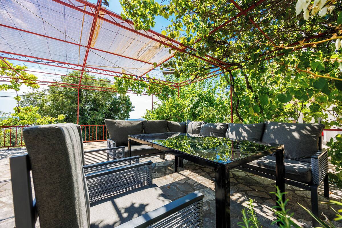Un área de estar al aire libre con mesa de vidrio, sillas de metal y un sofá bajo una estructura metálica roja.