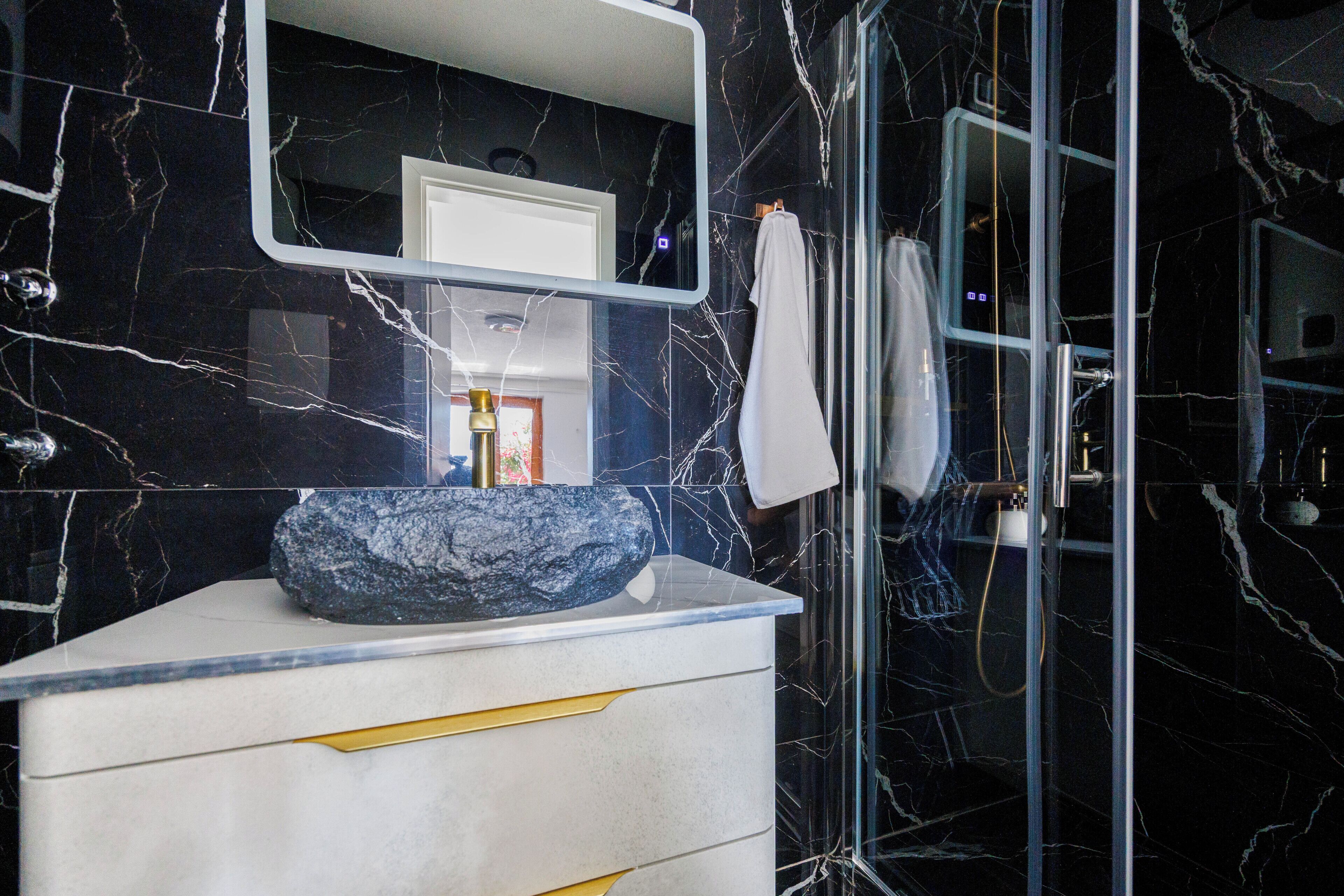A bathroom with a marble wall, a sink with a large stone basin, a mirror, and a glass shower enclosure.