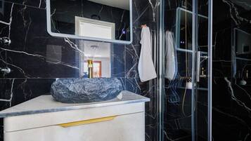 A bathroom with a marble wall, a sink with a large stone basin, a mirror, and a glass shower enclosure.