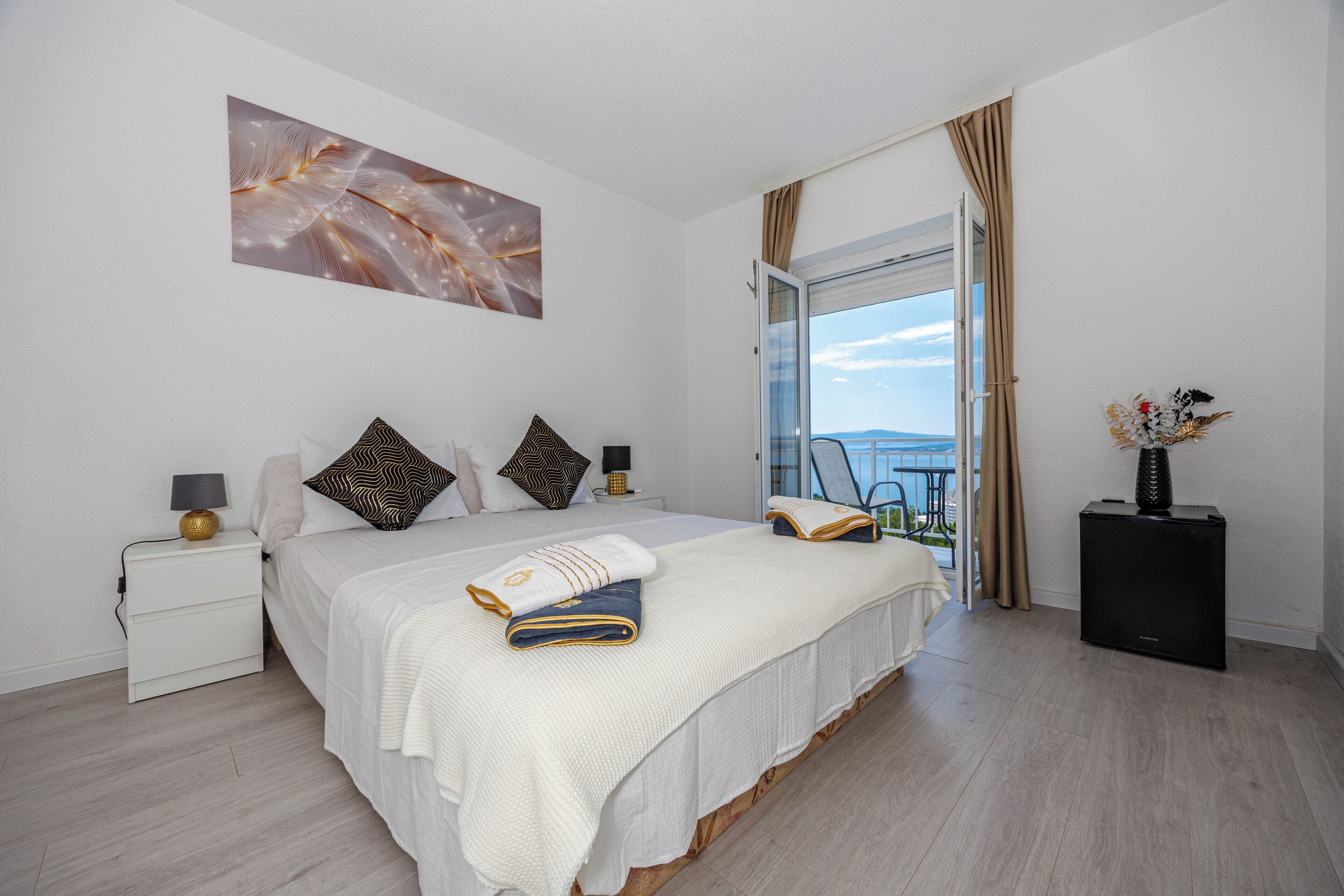 A modern hotel room with a large bed, a nightstand, a side table, and a view of the sea through an open door.