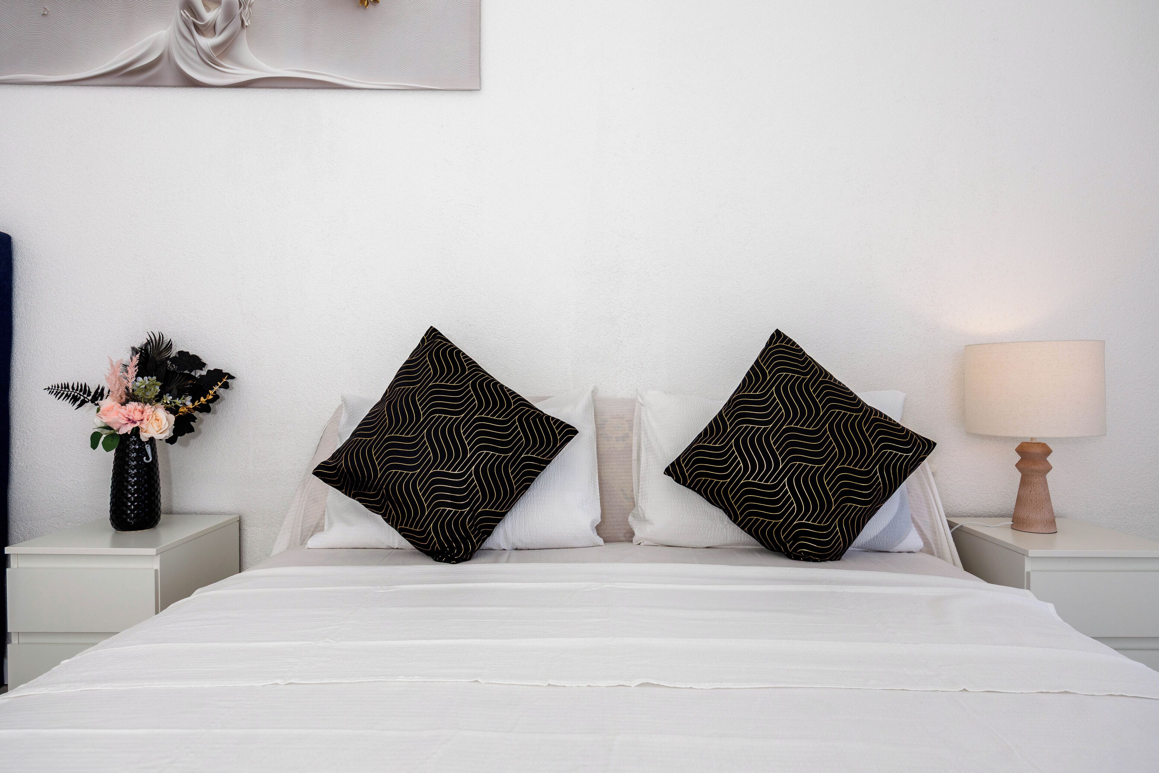 A neatly made bed with white bedding, two black patterned pillows, a vase with flowers, and a lamp on a bedside table.