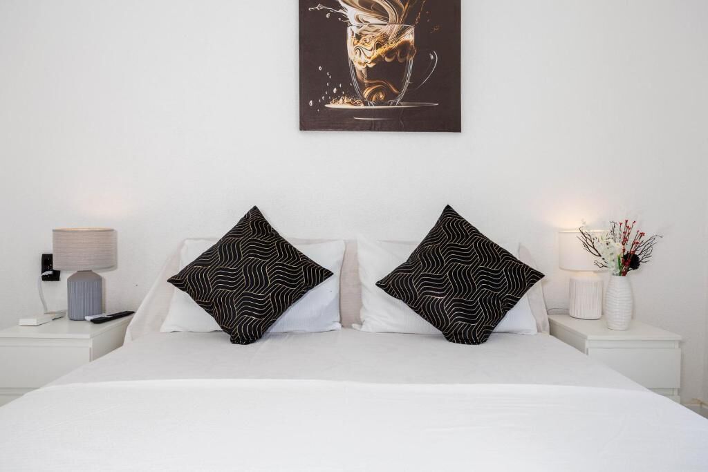 Un letto rifatto con due cuscini a motivi bianco e neri, un comodino bianco con una lampada e un'opera d'arte appesa al muro raffigurante una tazza da caffè.
