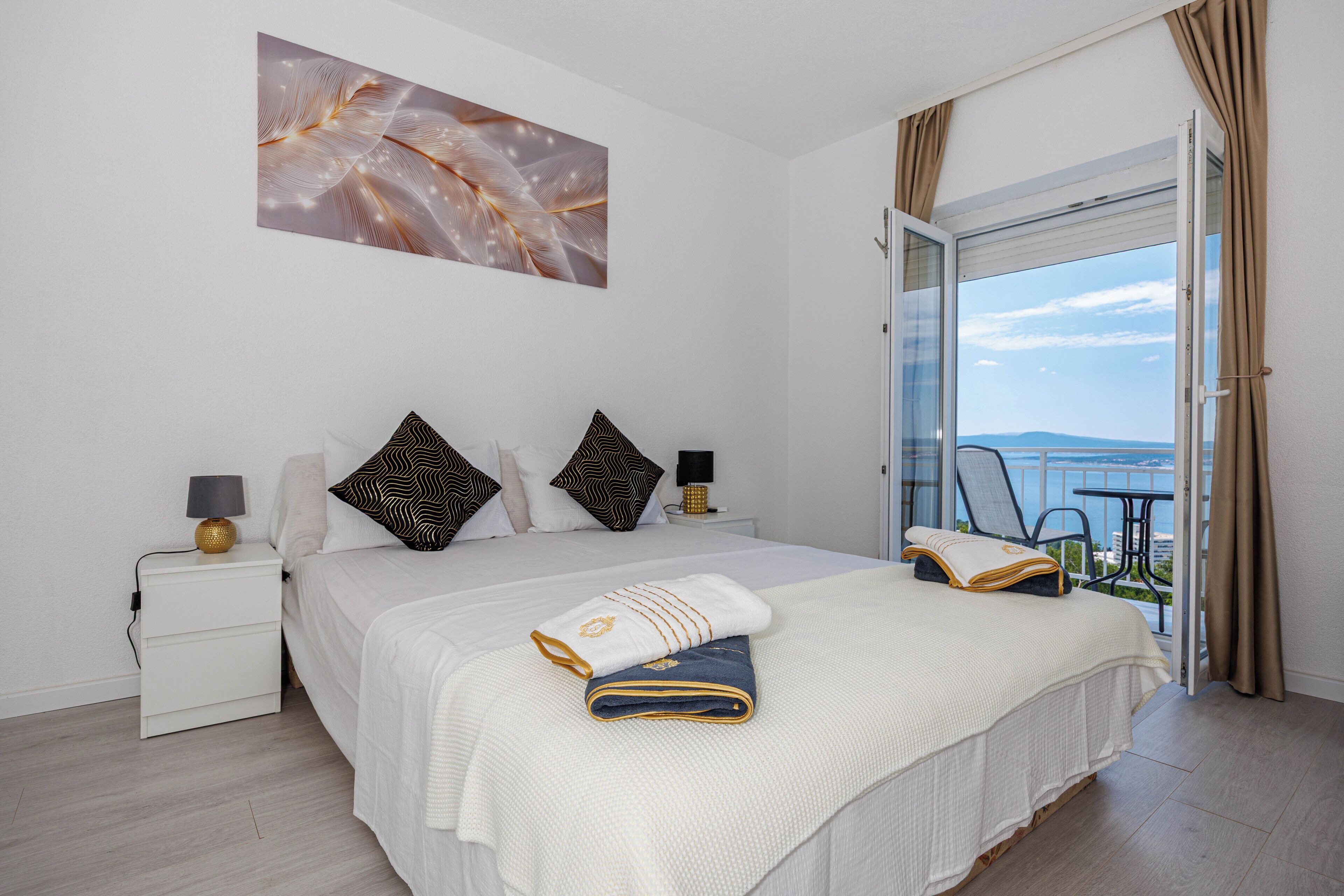 A bedroom with a bed, pillows, a nightstand, a reading lamp, a window with curtains, and a view of the sea.