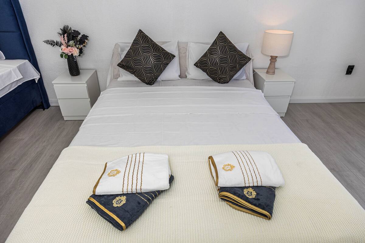 Una cama bien hecha con dos almohadas, una lámpara de noche y una mesita de noche con un florero.