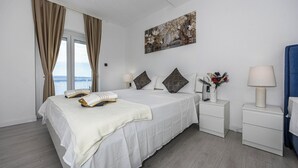 Un cuarto de hotel con una cama, dos mesitas de noche, un atril, un cuadro y vistas al mar.
