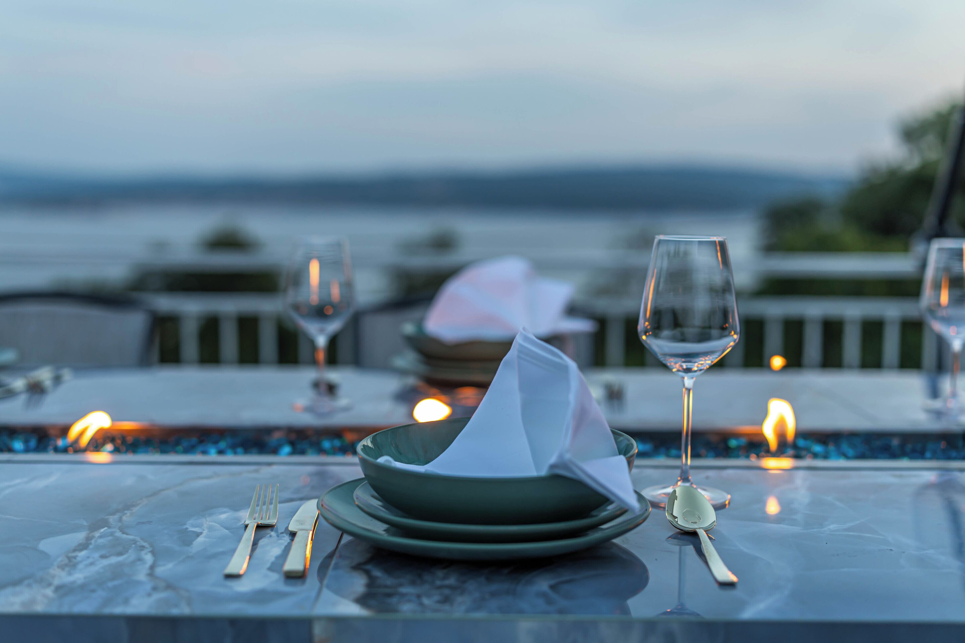 Mesa de comedor con platos, cubiertos y vasos, con vistas a un cuerpo de agua.