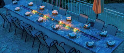 A long dining table set for a meal with chairs, a grill, and a decorative water feature.