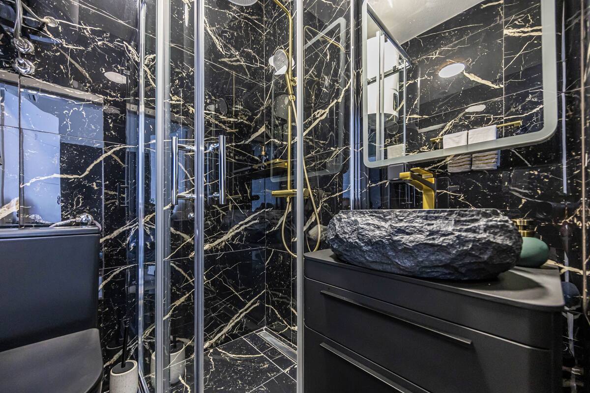 Un baño con ducha de vidrio, pared de azulejos de mármol, lavamanos con una gran roca y un espejo.