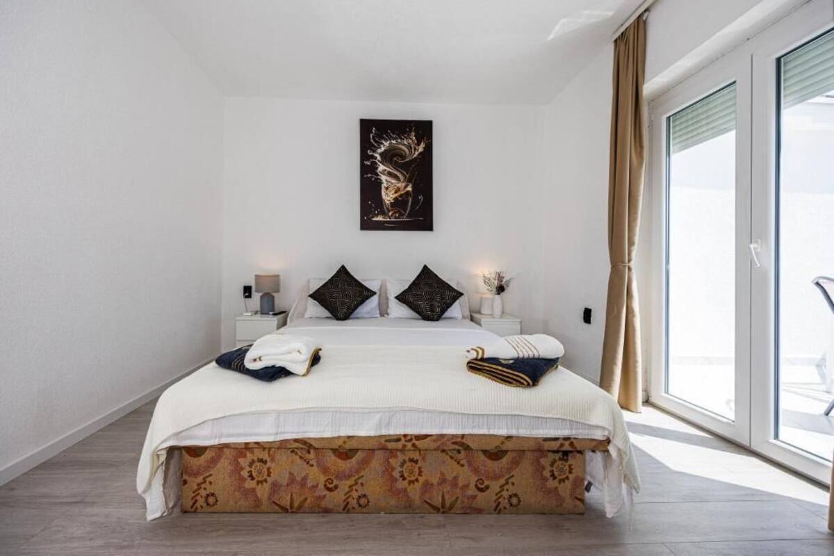 Un dormitorio con cama, almohadas, una mesita de noche, una obra de arte en la pared y una ventana con cortinas.