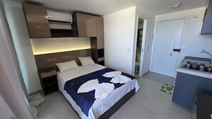 A hotel room with a bed, bedside table, built-in air conditioning unit, and a kitchenette with a sink and stove.