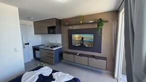 A modern bedroom with a bed, a small kitchenette, a TV mounted on the wall, and a window with curtains.