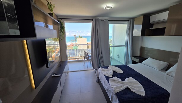 A modern hotel room with a bed, a TV, a balcony with a view of the city, and a small table with chairs.