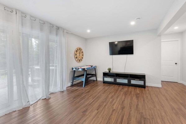 Una habitación con piso de madera, un televisor montado en la pared, un reloj y una ventana con cortinas traslúcidas.