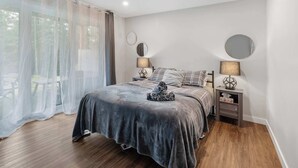 Un dormitorio con cama, dos mesitas de noche, ventana con cortinas traslúcidas y vista a zonas verdes.