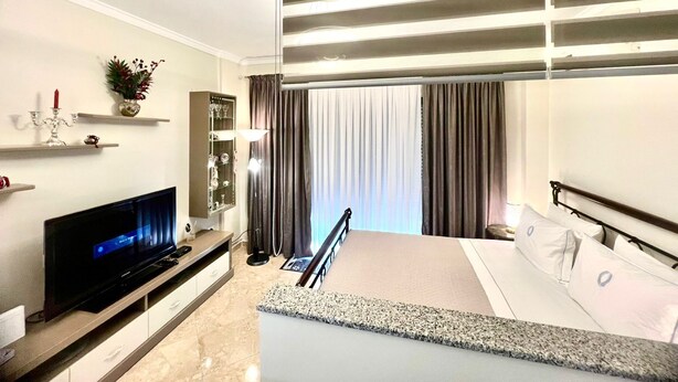 A modern hotel room with a flat-screen TV, a built-in shelf with decorative items, a large window with curtains, and a balcony with a railing. - Anna Seaview Cozy 1-Studio Apartment in charming Durres with Wi Fi, AC (Durres)
