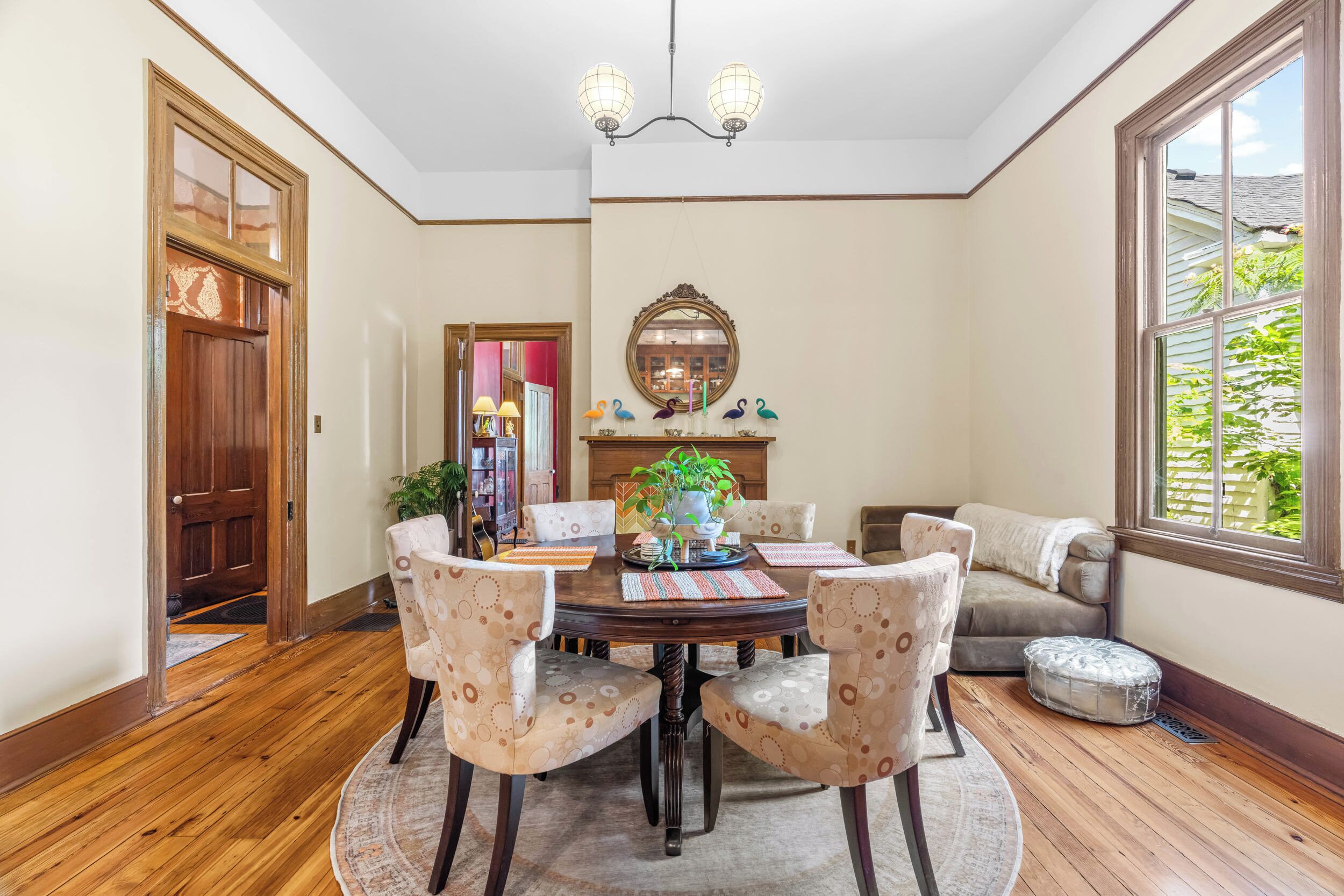 A dining area with a wooden table, four chairs, a chandelier, a mirror, and a window with a view of greenery.