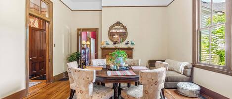 A dining area with a wooden table, four chairs, a chandelier, a mirror, and a window with a view of greenery.