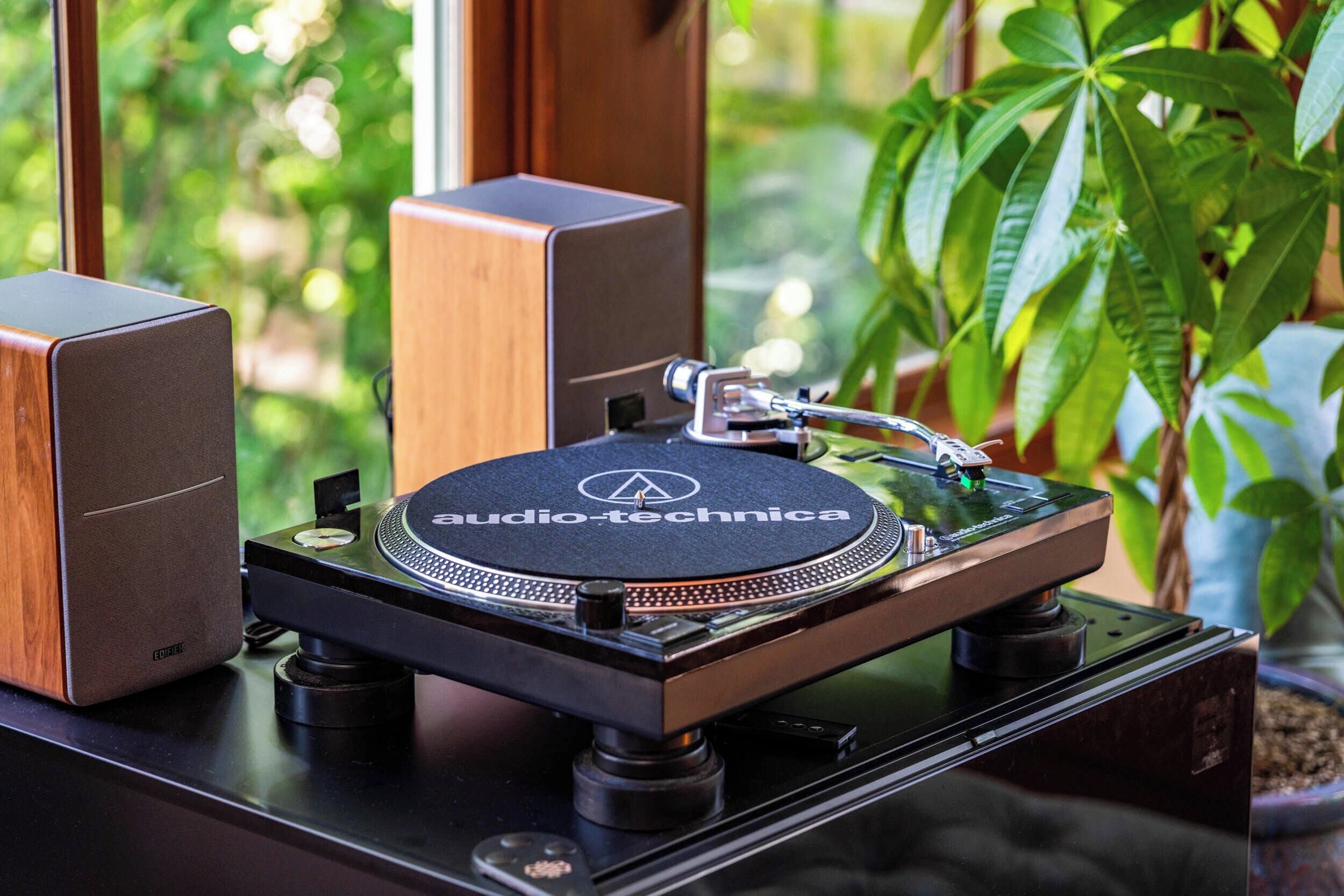 A turntable with a wooden cabinet and speakers, placed on a table near a window with a view of greenery.