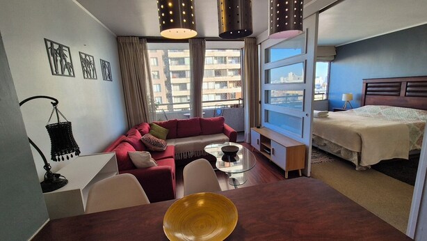 Habitación de hotel moderna con sofá rojo, cama, zona de comedor y grandes ventanales con vistas a la ciudad.