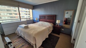 Un dormitorio con cama, mesita de noche con lámpara, ventana con vista a edificios y radiador.