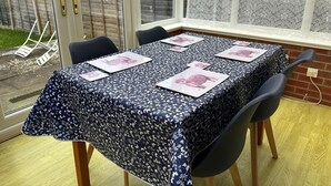 A conservatory with a table covered in a floral tablecloth, six chairs, and a view of a garden and houses through the glass doors. - Budget Carlisle Home for 4 | Parking & Garden (Carlisle)