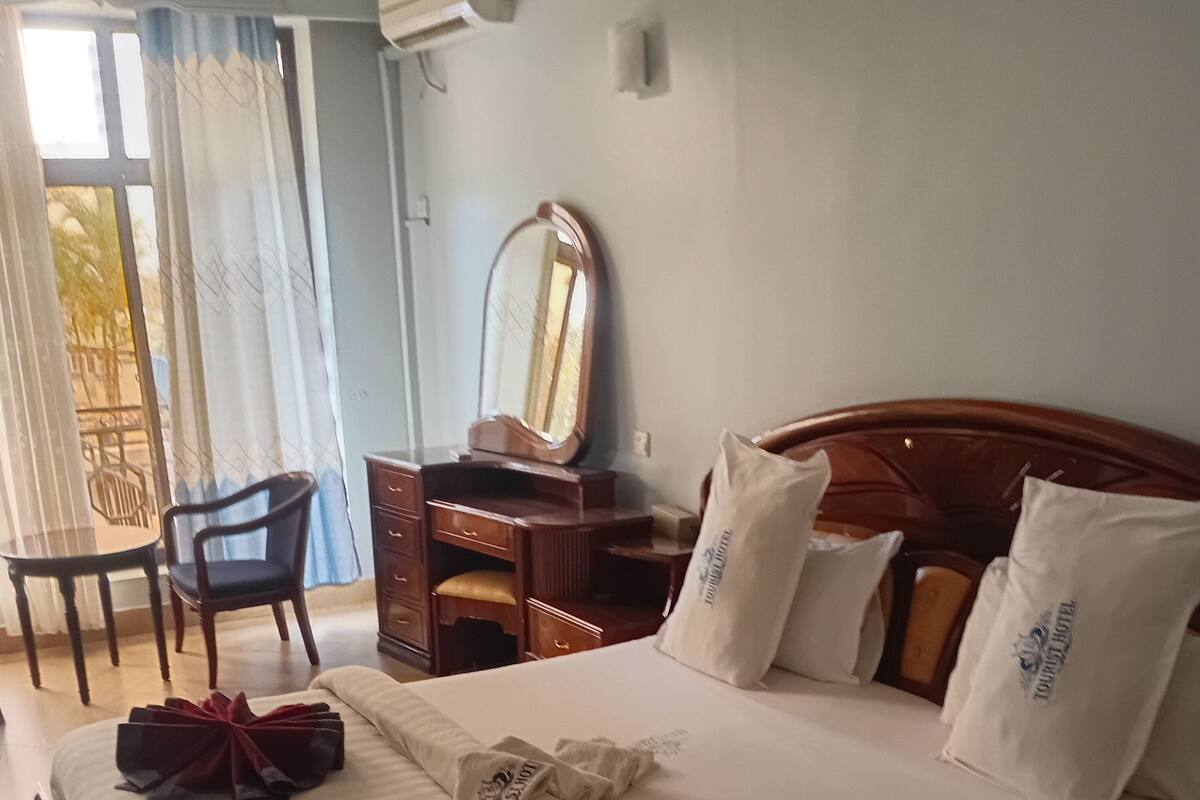 A hotel room with a bed, a wooden headboard, a desk with a mirror, a chair, and a patterned rug.