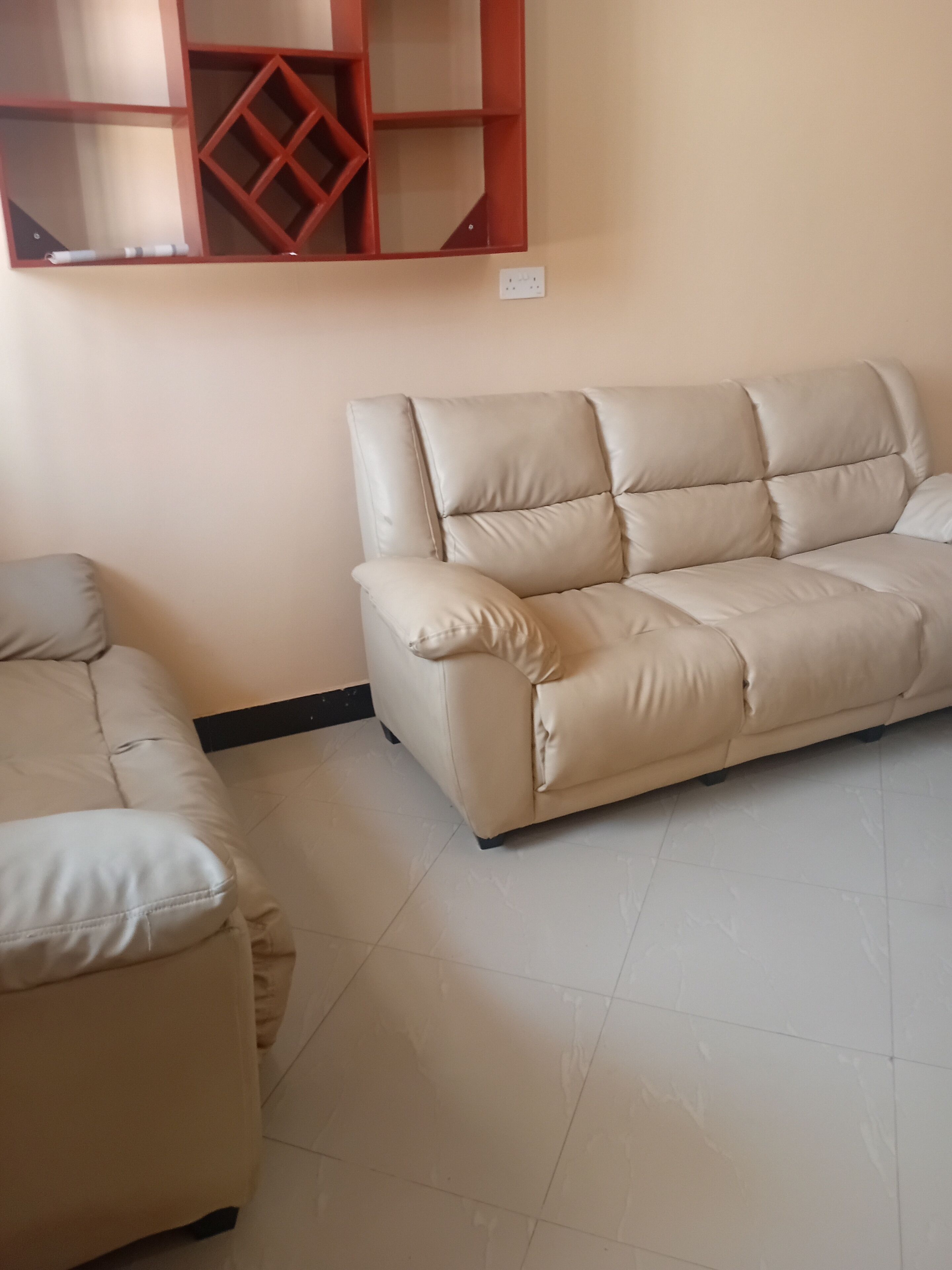 A beige sofa and a matching ottoman in a room with a tiled floor and a wall-mounted shelf unit.