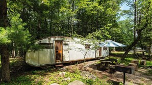 A vintage camper parked in a wooded area with picnic tables and a small shelter. - Outlier Inn Vintage Trailer Upstate Catskills Farm (Woodridge)