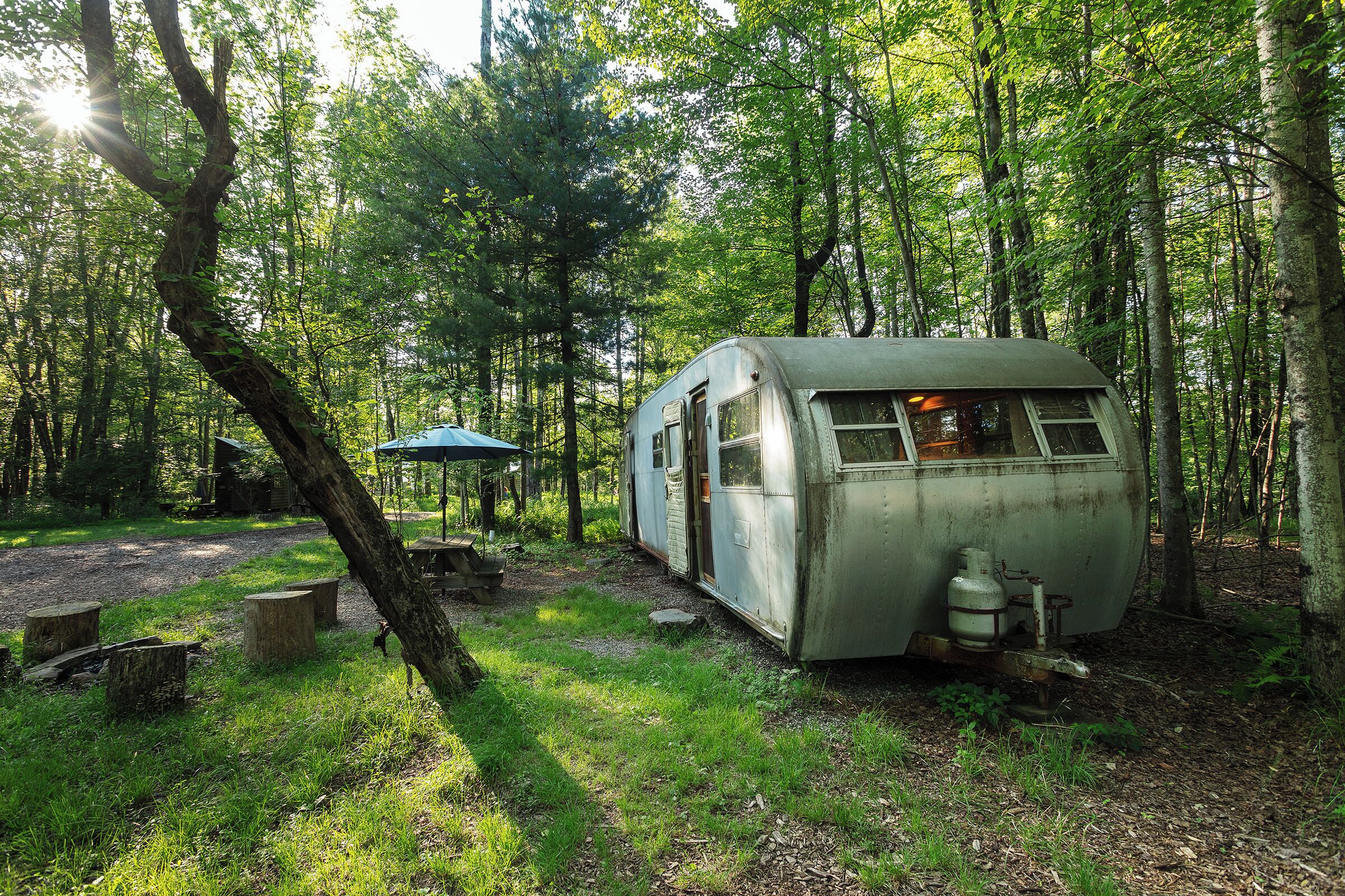 A vintage camper parked in a forested area with a picnic table and a blue umbrella nearby.