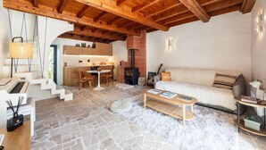 A cozy living area with a stone floor, wooden ceiling beams, a sofa, a coffee table, and a kitchen in the background.