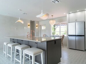 A modern kitchen with a large island, bar stools, stainless steel appliances, pendant lighting, and a view to the outdoors.