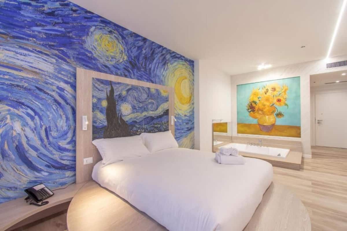 Habitación de hotel con cama, un cuadro de un cielo nocturno, un cuadro de girasoles y un baño con lavabo y espejo.
