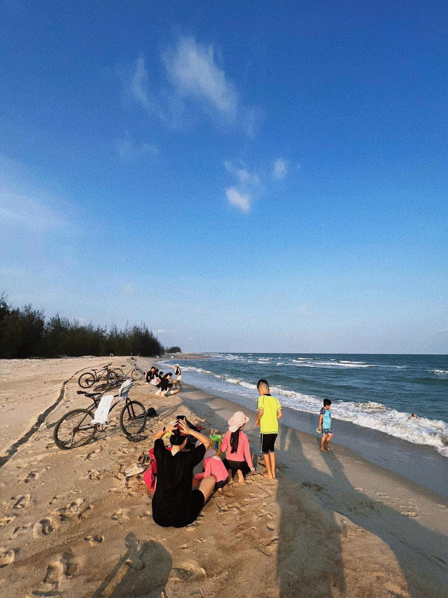 Uma praia com bicicletas estacionadas na areia, pessoas relaxando e o oceano ao fundo.