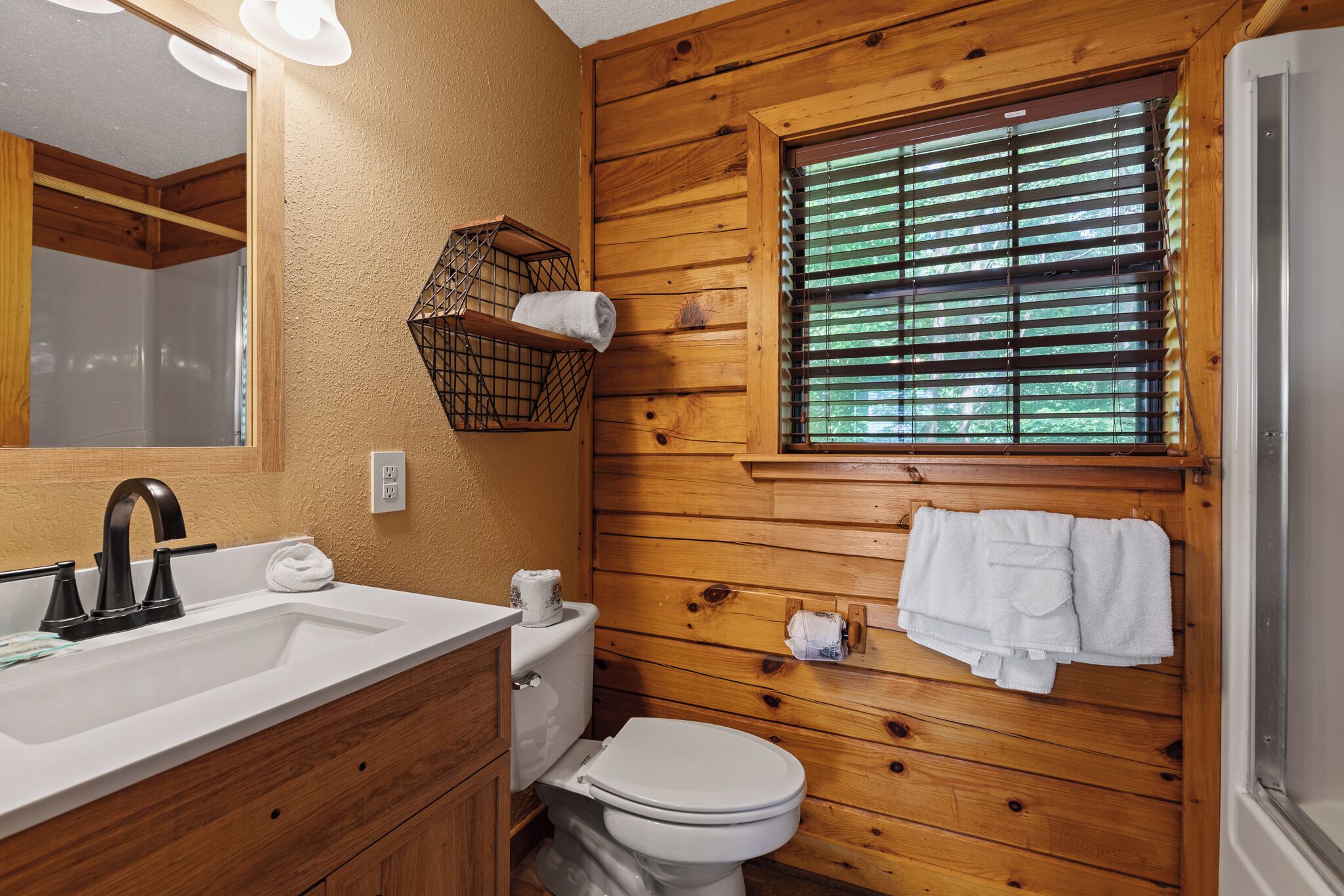 A wooden-paneled bathroom with a sink, toilet, and window with blinds.