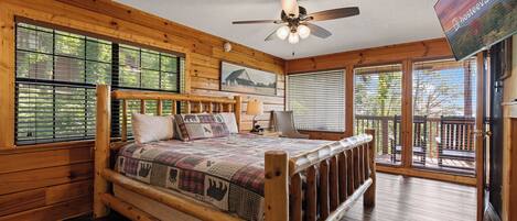 A wooden cabin bedroom with a bed, a ceiling fan, a TV, and large windows.