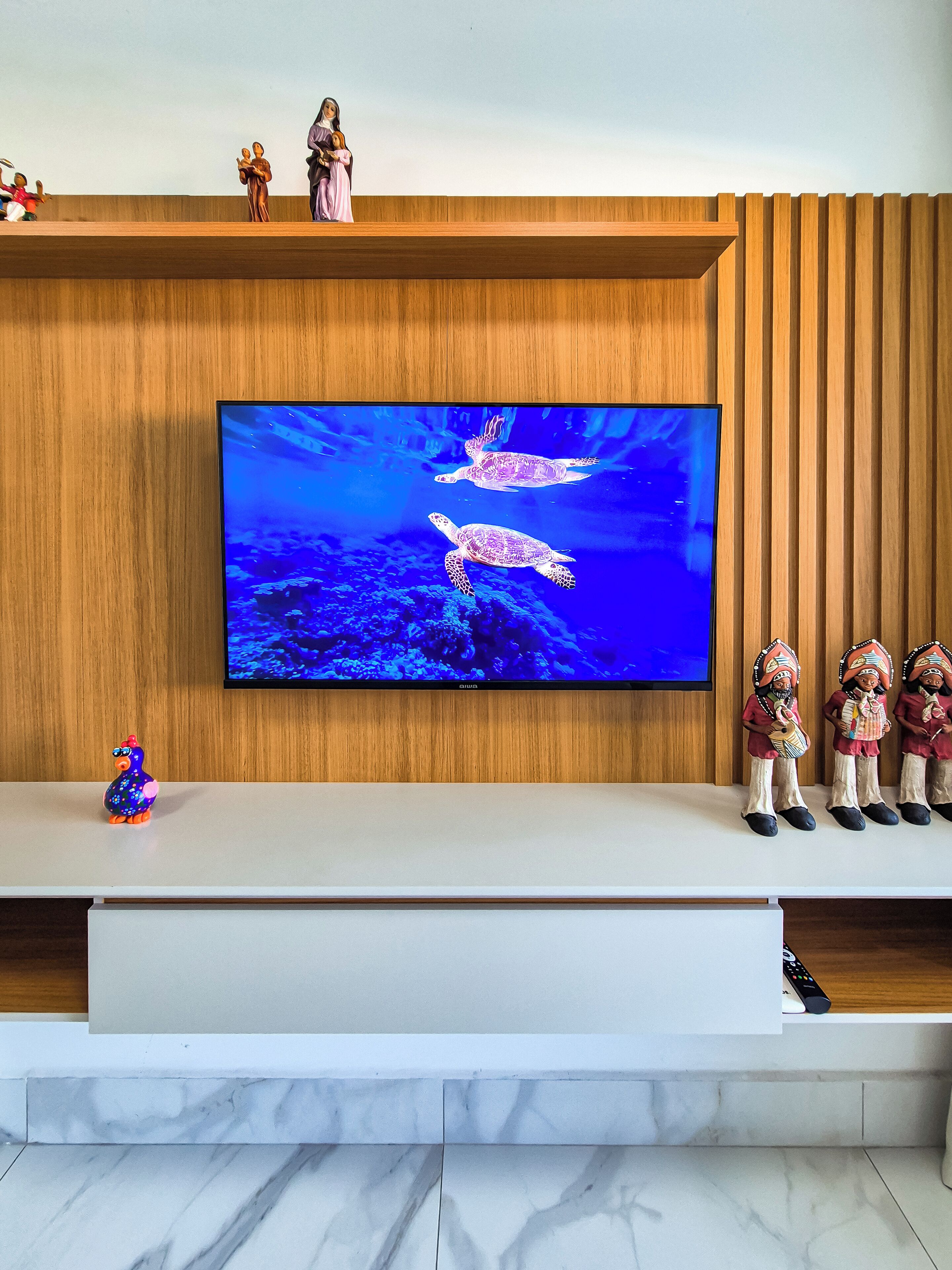 A flat-screen TV mounted on a wooden wall, with a shelf above displaying figurines and a small toy bird.