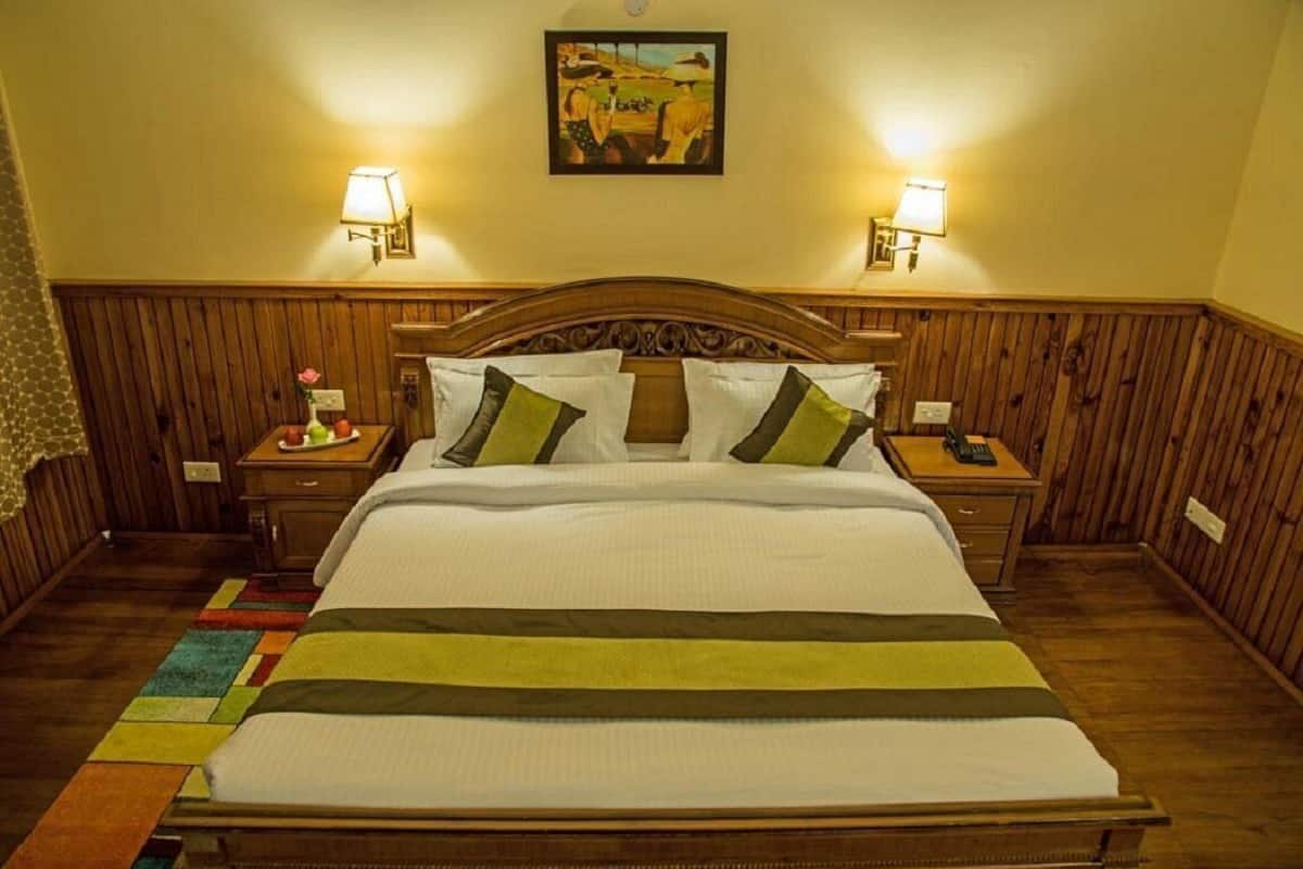 Hotelkamer met houten hoofdeinde, twee bedlampen, een bed met wit beddengoed en groene kussens, een kleurrijk tapijt en een schilderij aan de muur.