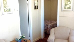 Ein Zimmer mit zwei beigen Sesseln, einem kleinen Holztisch mit Blumenvase und einem Bett mit floralem Überwurf. Eine Tür führt in einen anderen Raum.