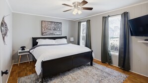 Un dormitorio con cama, ventilador de techo, mesita de noche, televisor y ventana con cortinas.