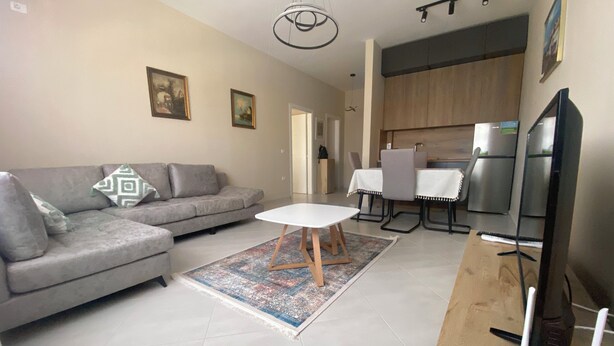 A modern living room with a grey sofa, a small white table, and a kitchen area with a fridge and a dining table.