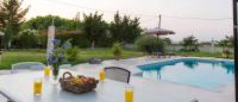 An outdoor pool area with a table set for dining, surrounded by greenery and a clear sky.
