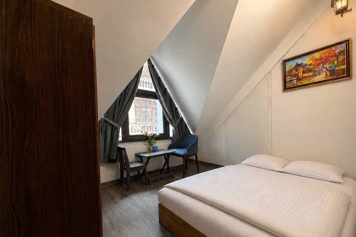 A bedroom with a bed, a desk, and a chair, a window with curtains, and a painting on the wall.